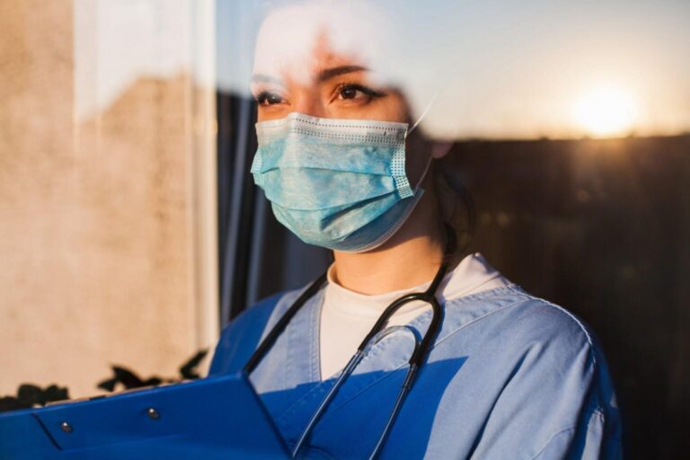 Young sad female caucasian UK US GP EMS doctor carer looking through ICU window,fear uncertainty in eyes,wearing face mask gazing at sun,hope faith in overcoming Coronavirus COVID-19 pandemic crisis